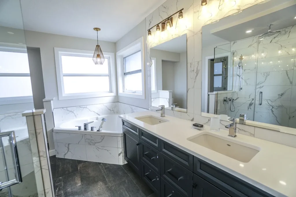 Modern bathroom with white marble walls, double sinks with a large mirror, dark cabinets, a glass shower, and a bathtub under two windows. Warm lighting above the mirror and pendant light over the tub.