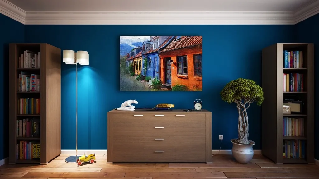 A cozy living room with blue walls features a painting of colorful houses. A wooden cabinet with drawers is flanked by modern floor lamps, a potted plant, and bookshelves. A toy car and alarm clock sit on the cabinet.