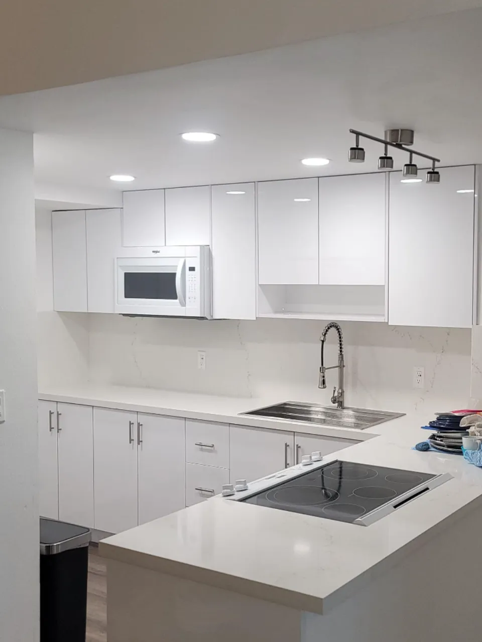 Modern kitchen with white cabinetry and countertops. Features an electric stovetop on the island, a stainless steel sink with a flexible faucet, an overhead microwave, and recessed lighting. A black trash bin is visible to the left.